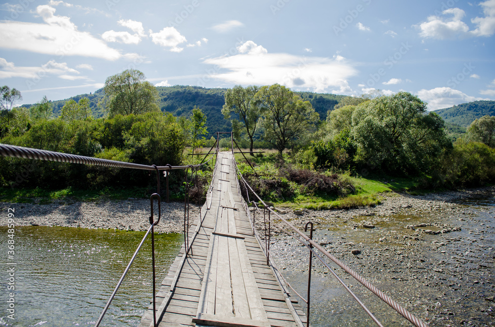 View of a rocky shallow mountain river from a tall old wooden suspension bridge in sunlight