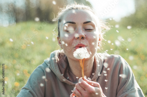 Young spring fashion woman blowing dandelion in spring garden. Springtime. Trendy girl at sunset in spring landscape background. Allergic to pollen of flowers. Spring allergy