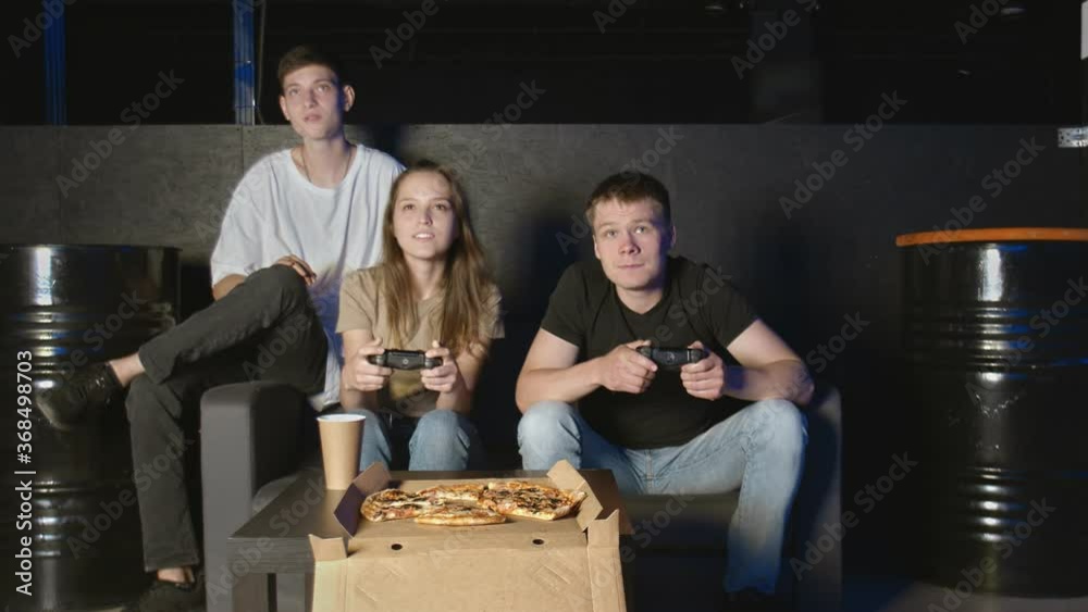 Friends play on the game console. They are sitting on the couch and holding the joysticks. One man winning another losing Friends play on the game console. They are sitting on the couch and holding the joysticks. One man winning another losing