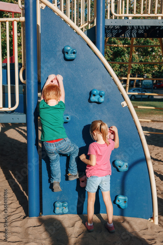 Little preschool boy and girl climbing rock wall at playground outside on summer day. Happy childhood lifestyle concept. Seasonal outdoors activity for kids. View from back.