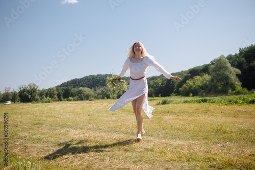 Young white woman with long blondy hair dressed in a white dress is dancing in a field with a wreath