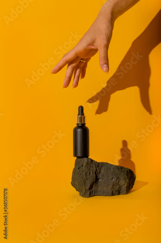 A cosmetic bottle with a pipette balances on a gray stone against a yellow background. A woman's hand gracefully reaches for the bottle. Hard shadows.