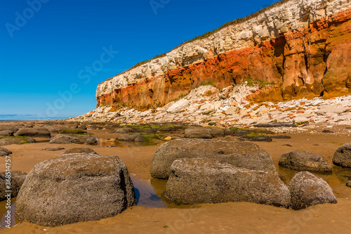 Barnacle encrusted rocks, rockfalls of varying colour and partially reflecting rock pools at the base of the white, red and orange stratified cliffs at Old Hunstanton, Norfolk, UK