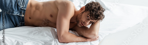 Panoramic shot of handsome muscular man looking at camera on bed isolated on white