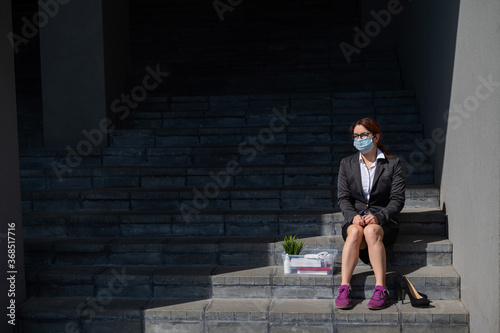 Dismissed woman with a box of personal belongings sits on the stairs on the outside. Female employee in medical mask fired due to financial crisis in coronovirus. Unemployment.