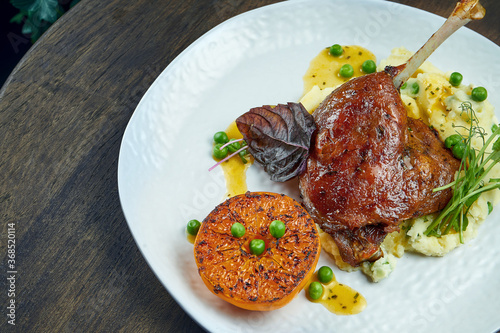 Appetizing confit duck leg with grilled orange and mashed potato garnish on a white plate on a wooden background