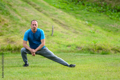 Always in a great shape. Sportsman is stretching standing over green natural background. Full length of strong young muscular man in comfortable sportwear stretching his arms and legs