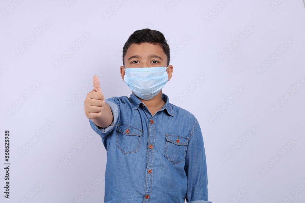 Cute Indian boy wearing surgical mask showing thumbs up or all done or like hand gesture. Isolated over white background.
Cute Indian boy wearing surgical mask showing thumbs up or all done or like hand gesture. Isolated over white background.