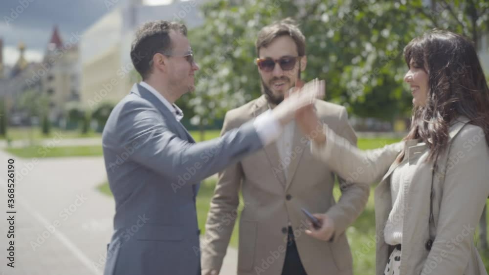 Portrait of confident elegant businessman in formal suit and sunglasses meeting friends on urban street and leaving with them. Young positive Caucasian guy greeting man and woman outdoors. Portrait of confident elegant businessman in formal suit and sunglasses meeting friends on urban street and leaving with them. Young positive Caucasian guy greeting man and woman outdoors.