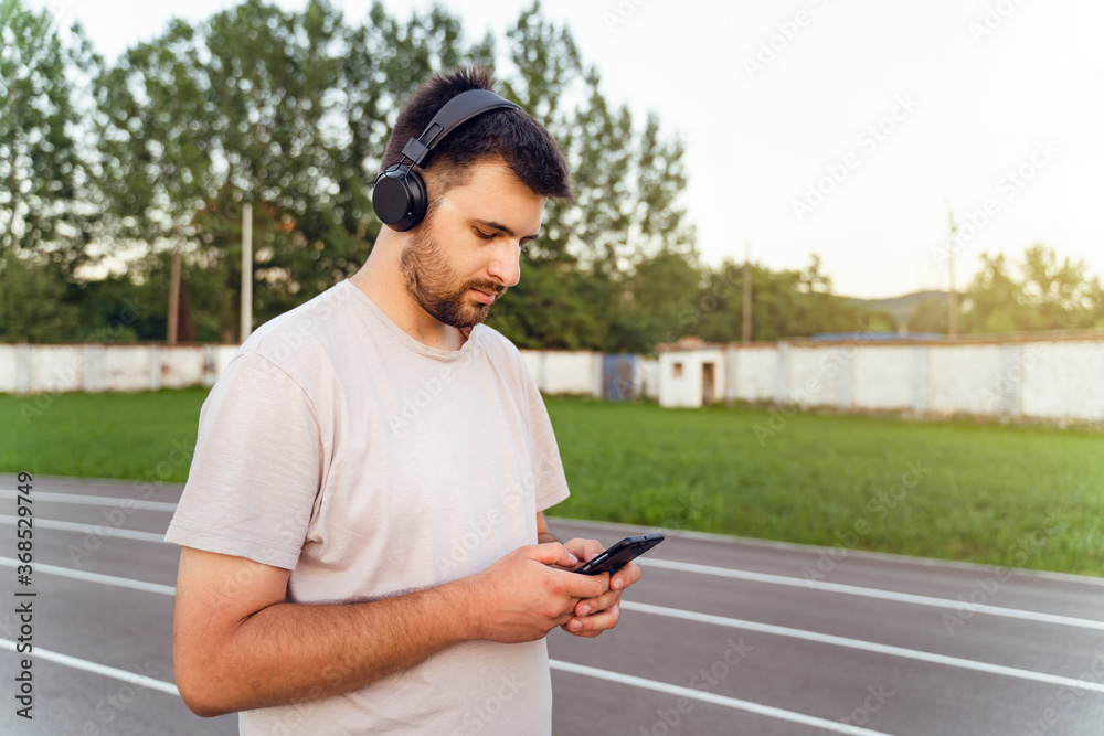 Fototapeta premium Man standing on the running track in day with smart phone and earphones - Adult caucasian male using mobile application to monitor training stats make a call send message - real people focus concept