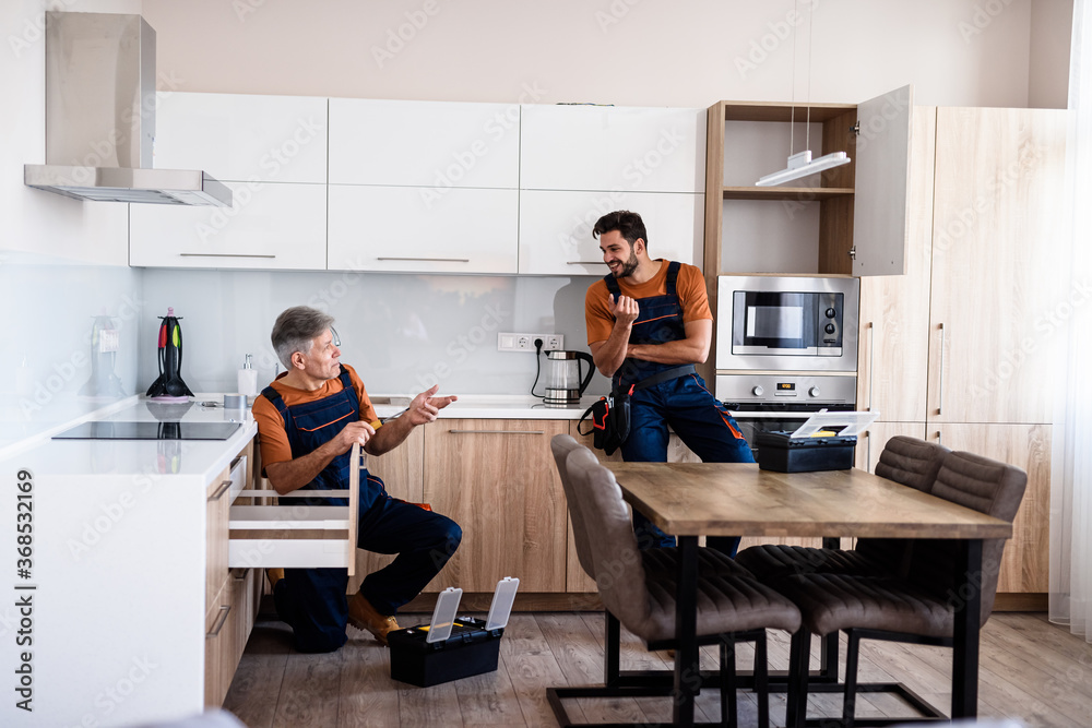 Full length shot of two handymen, workers in uniform talking while assembling kitchen cabinet using screwdriver indoors. Furniture repair and assembly concept Full length shot of two handymen, workers in uniform talking while assembling kitchen cabinet using screwdriver indoors. Furniture repair and assembly concept