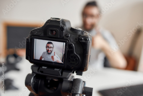 Young male blogger showing thumbs up at camera while recording video blog or vlog at home studio. Focus on camera screen