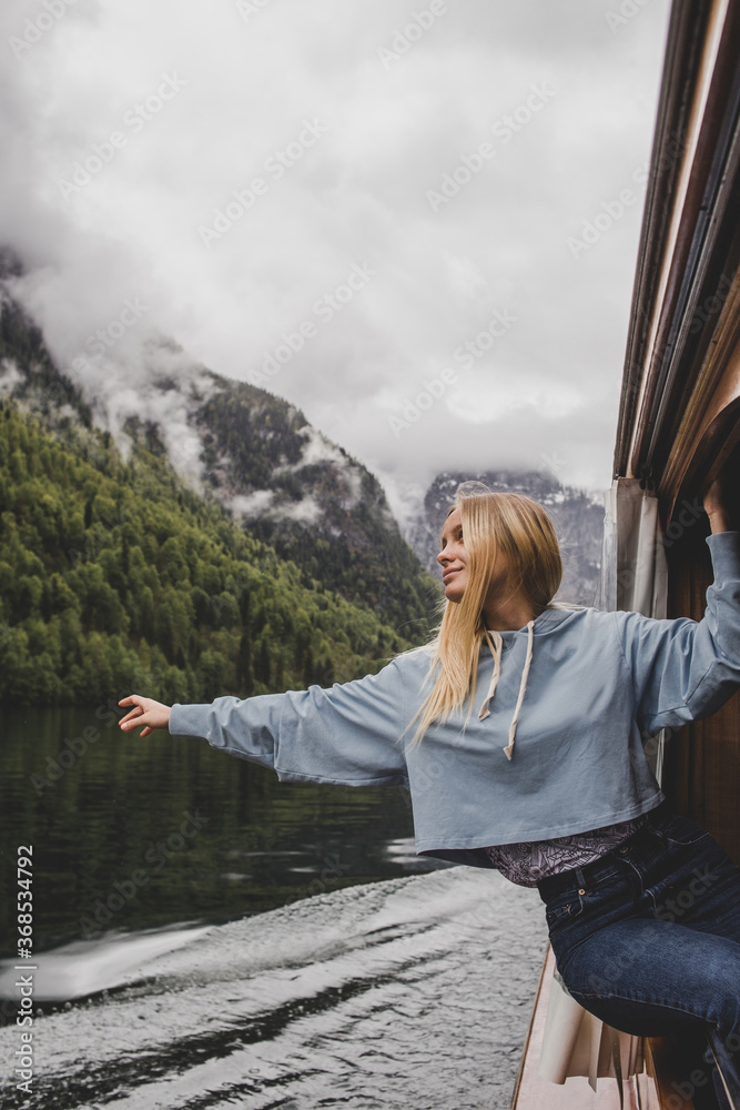 Konigssee. Bavaria. Germany. Blonde caucasian girl feels freedom from the window of the wooden boat on background of lake, forest & rock mountain in clouds in rainy spring day