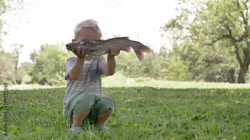 Child (little boy) holding a big fish (catfish) in his hand. Outdoors activities, fishing, catching fish concept.  Daytime, summer 
