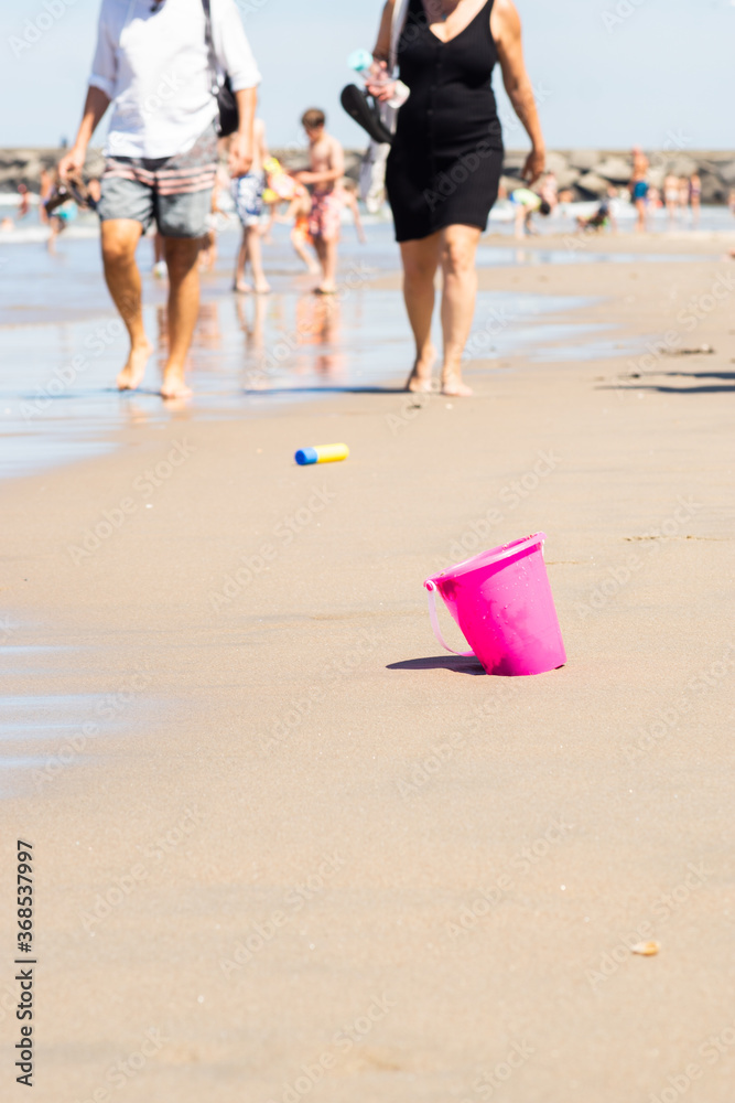 Plastic pink toy bucket left alone on the beach sand near ocean water, blurred people on background - concept of vacation safety, water drowning, safe kids, also plastic pollution during summer