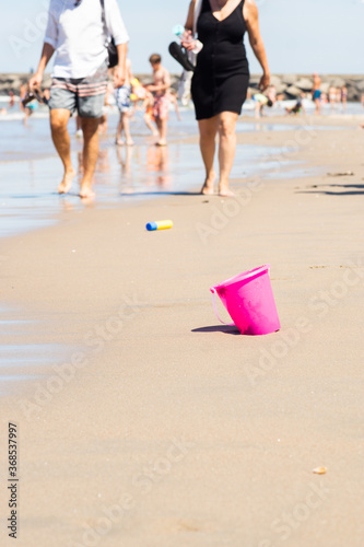 Obraz na płótnie Plastic pink toy bucket left alone on the beach sand near ocean water, blurred people on background - concept of vacation safety, water drowning, safe kids, also plastic pollution during summer w salonie
