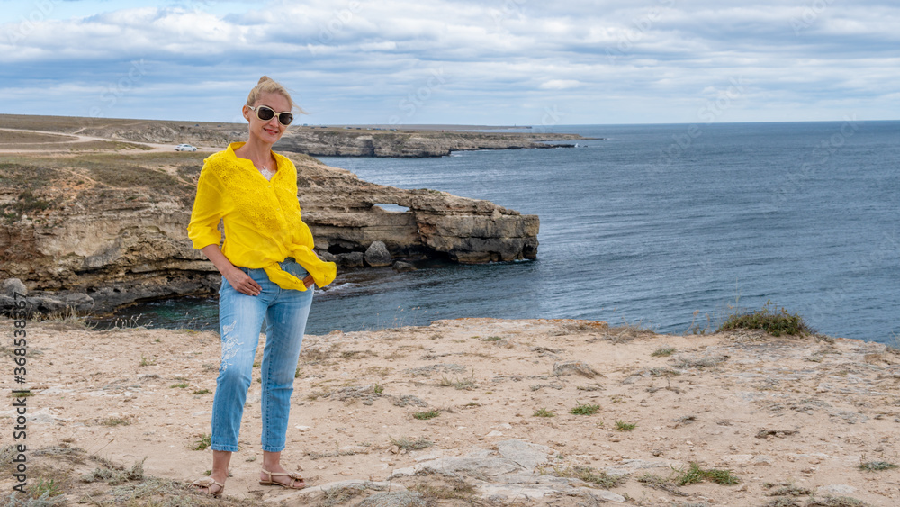 Girl stands on against rocks resembling a crocodile at sea, yellow shirt