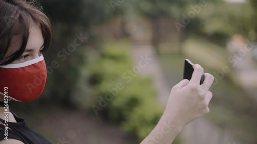 Young girl wearing a red face mask taking a selfie photo using smartphone walking in the park during Covid-19 Crisis