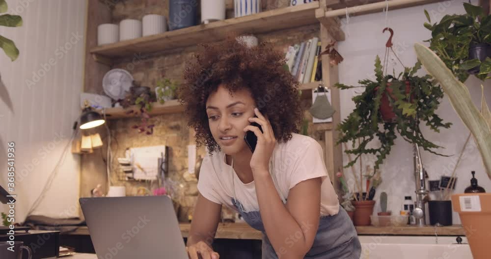 Young adult Afro American female small business owner working on laptop in store and speaking to client on phone