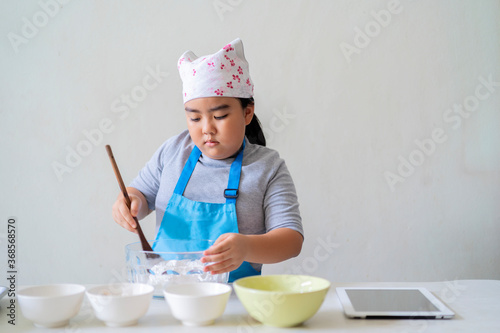 A cute Asian kid is learning sweets, she is pouring the ingredients together. Inside your home kitchen, An Asian girl is learning to cook