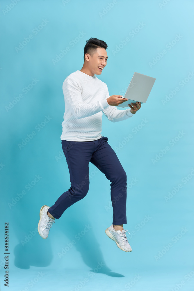 Full length portrait of happy man jumping and holding laptop isolated over blue background Full length portrait of happy man jumping and holding laptop isolated over blue background