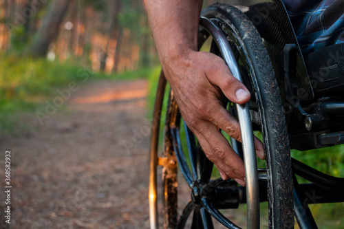 A disabled person is sitting in a wheelchair. He holds his hands on the steering wheel, walks in the park