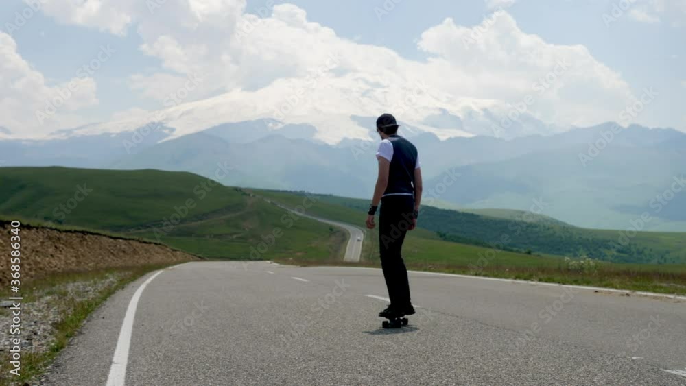 guy in summer in black clothes descends from the mountain on a skateboard against the background of the snow-covered mountain elbrus guy in summer in black clothes descends from the mountain on a skateboard against the background of the snow-covered mountain elbrus