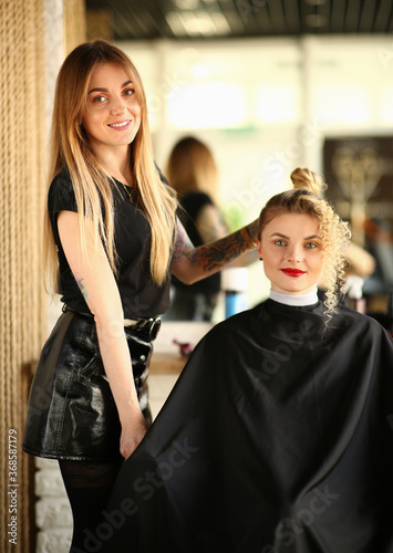 Blonde Hairstylist and Curly Hair Female Client. Beautiful Hairdresser Styling Wavy Strand and Bun for Young Woman. Two Girls in Beauty Salon. Styling Hair Curls for Customer Looking at Camera Shot