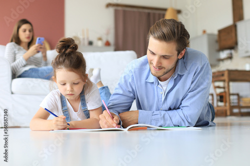Happy father smilling daughter lying on warm floor enjoying creative activity, drawing pencils coloring pictures in albums, mother resting on couch, family spend free time together.
