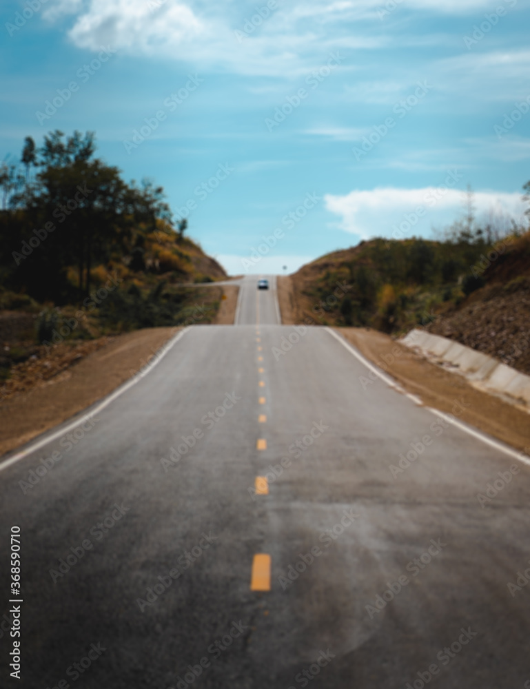 Blur background of gray asphalt road in a peaceful rural area on a sunny day and clear blue sky clouds. There are white border and yellow central line. The roadside is brown, grass, and green trees. Blur background of gray asphalt road in a peaceful rural area on a sunny day and clear blue sky clouds. There are white border and yellow central line. The roadside is brown, grass, and green trees.