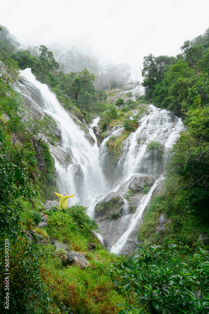 A woman wearing a yellow raincoat, standing with her back turned. Blur backdrop is a waterfall in the rainy season, Thailand. Feeling fresh, relaxed, and freedom. Ideas for adventure nature tourism.