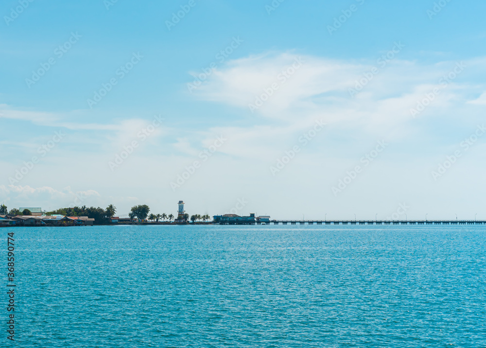 A photograph panorama on the boat of the sea view in the morning. The backdrop has a port, a citadel, coconut palms, and a seaside village in Thailand. There are clear skies and bright pastel blue.