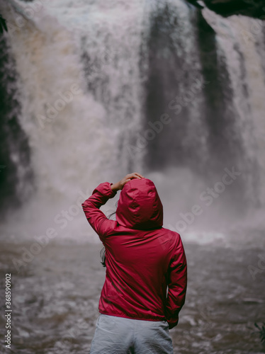 A woman wearing a red hood, standing with her back turned. Blur backdrop is wonderful waterfall in the rainy season, Thailand. Feeling fresh, relaxed, and freedom. Ideas for adventure nature tourism. © Pang wrp