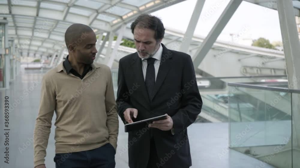 Male business partners using tablet, discussing content on screen while walking at train station. Dolly, medium shot, front view. Communication concept Male business partners using tablet, discussing content on screen while walking at train station. Dolly, medium shot, front view. Communication concept