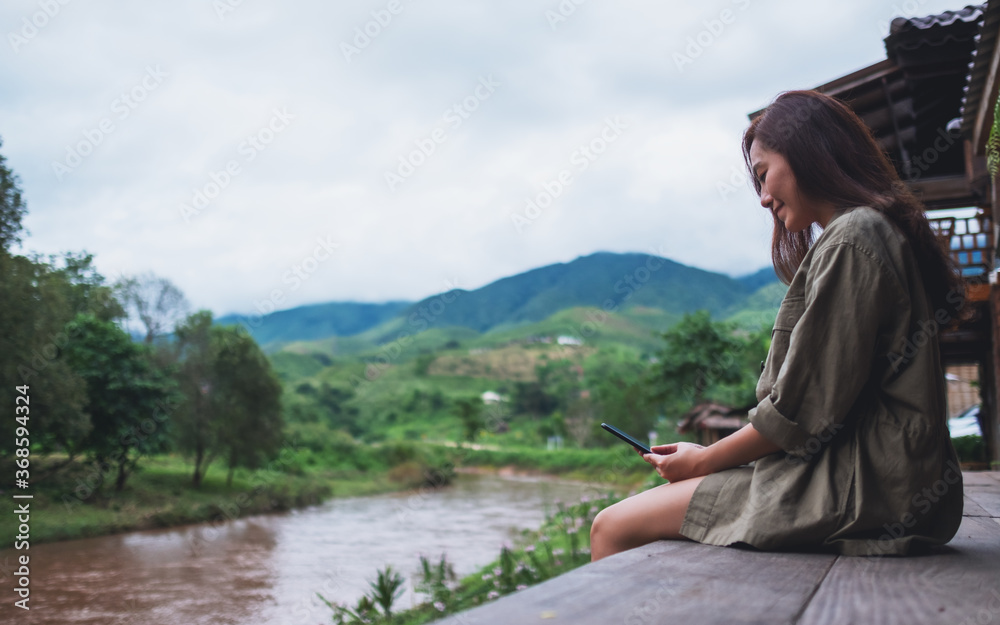 A beautiful asian woman using mobile phone while sitting by the river with mountains and nature background