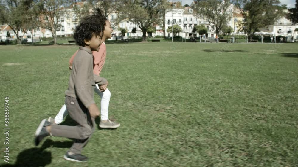 Happy mixed-race children running on grass, holding hands and smiling in slow motion. Buildings on background. Tracking camera. Side view. Childhood, fun and summer weekend concept Happy mixed-race children running on grass, holding hands and smiling in slow motion. Buildings on background. Tracking camera. Side view. Childhood, fun and summer weekend concept