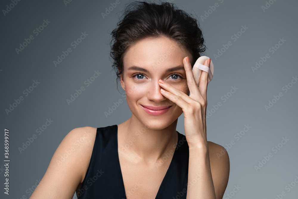 Beautiful woman applying dry powder, using cosmetic cushion on her facial skin. Photo of woman with perfect makeup on gray background. Beauty concept Beautiful woman applying dry powder, using cosmetic cushion on her facial skin. Photo of woman with perfect makeup on gray background. Beauty concept