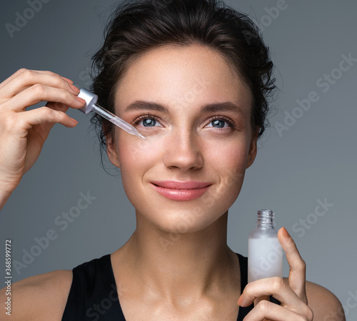 Woman applying hyaluronic serum on fer face with pipette. Photo of attractive woman with perfect makeup on gray background. Beauty concept