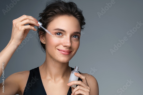 Woman applying  hyaluronic serum on fer face with pipette. Photo of attractive woman with perfect makeup on gray background. Beauty concept photo