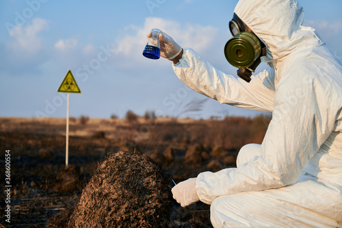 Ecologist in suit, gas mask holding test tube with blue liquid while studying burnt grass and soil on scorched territory with biohazard sign. Environmentalist doing laboratory test in field after fire