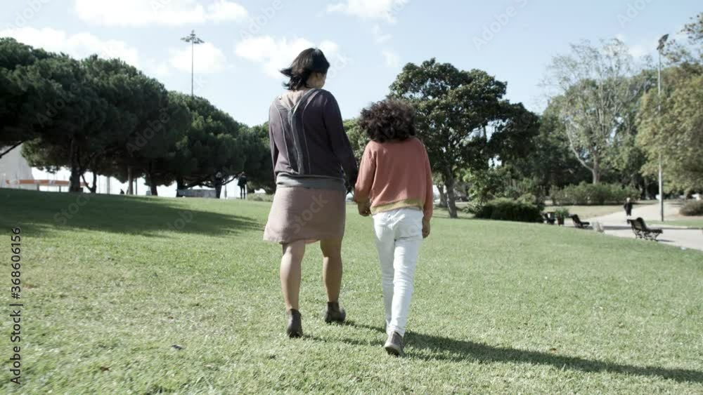 Asian mom holding mixed-race cute daughter hand and walking on meadow in windy day. Back view of little girl pacing with mother. Sky, bench and trees on background. Family, holiday and weekend concept Asian mom holding mixed-race cute daughter hand and walking on meadow in windy day. Back view of little girl pacing with mother. Sky, bench and trees on background. Family, holiday and weekend concept