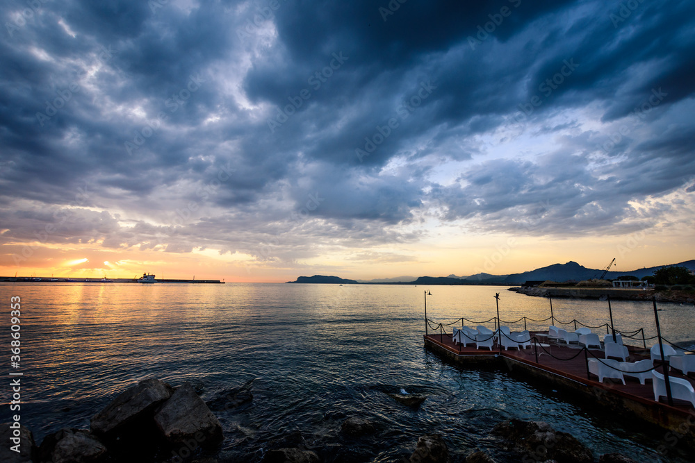Fototapeta premium Landscape of a port at sunrise, with tables and chairs in the foreground and the silhouette of a ship in the background
