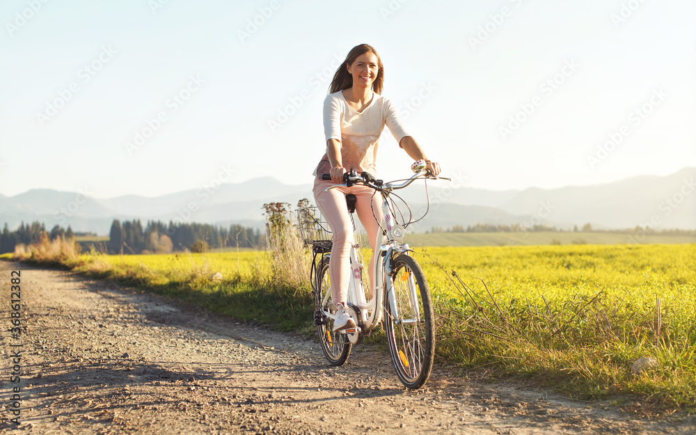 Young woman riding bike on dusty country road, smiling, blurred sun lit meadow behind her