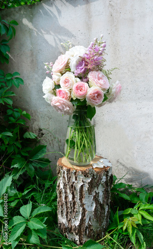 A romantic photo with a bouquet of delicate pink English roses standing in a vase on a birch tree stump. Romantic bouquet on the background of a concrete wall entwined with virginia creeper.
