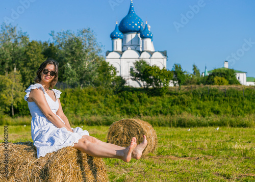 A young girl is resting in a field on a roll of hay, near a Christian church.
