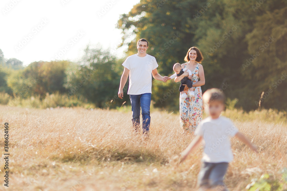 Fototapeta premium Family with children walking outdoors in summer field at sunset. Father, mother and two children sons having fun in summer field. People, family day and lifestyle concept