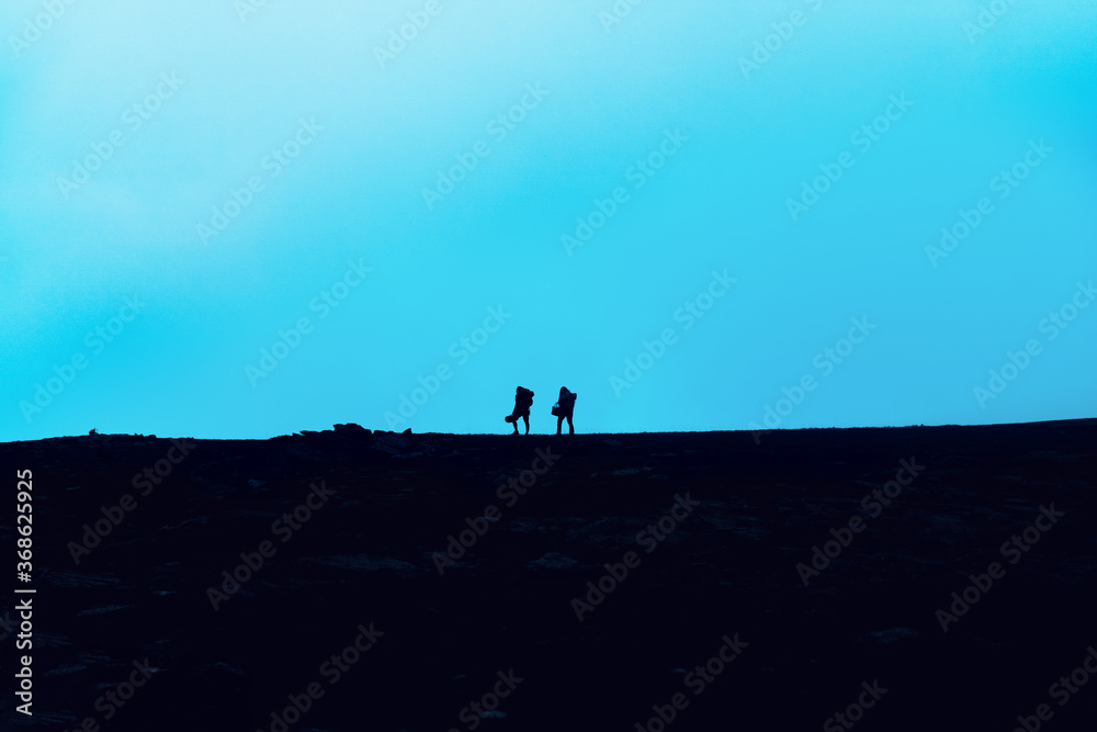 Travelers walk along a trail in the mountains. Silhouettes of two adventure hikers with backpacks walk in the mountains at dusk.