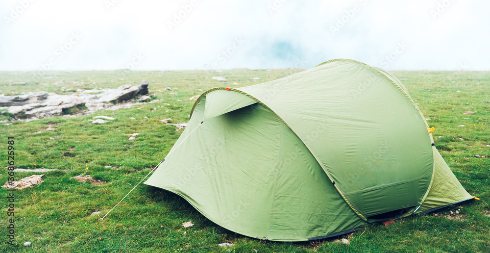 Tent in the wilderness. A single green camping tent in a grassy meadow with fog on the mountain. Trekking, camping, hiking and wild life concept. Real Outdoor Adventure.