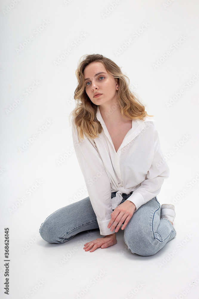 portrait of young pensive caucasian woman posing in shirt and blue jeans, sitting on white studio floor on her knees. model tests of pretty girl in basic clothes on cyclorama. attractive female poses