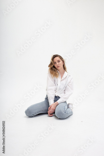 portrait of young pensive caucasian woman posing in shirt and blue jeans, sitting on white studio floor on her knees. model tests of pretty girl in basic clothes on cyclorama. attractive female poses